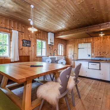 Ein Essbereich mit großem Holztisch, weißen Stühlen und einem Holzofen bei der Talsenalm